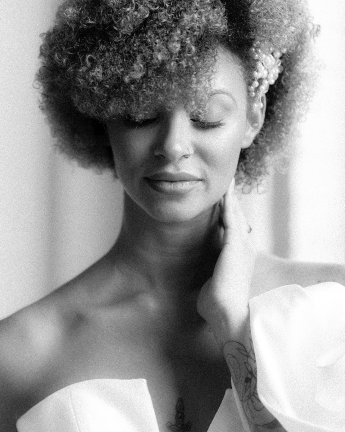 Black and white portrait of a woman with curly hair and closed eyes, touching her neck, wearing a white off-shoulder dress and a beaded hair accessory.