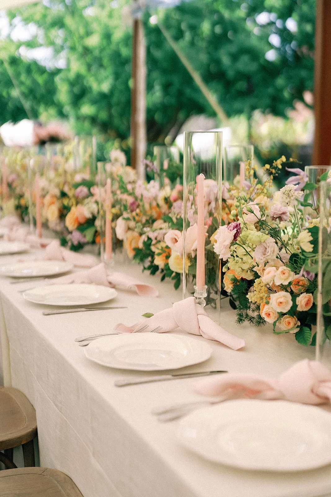 A long dining table decorated with pastel-colored flowers, pink candles in glass holders, with plates, silverware, and pink napkins on a white tablecloth. There are outdoor trees in the background.