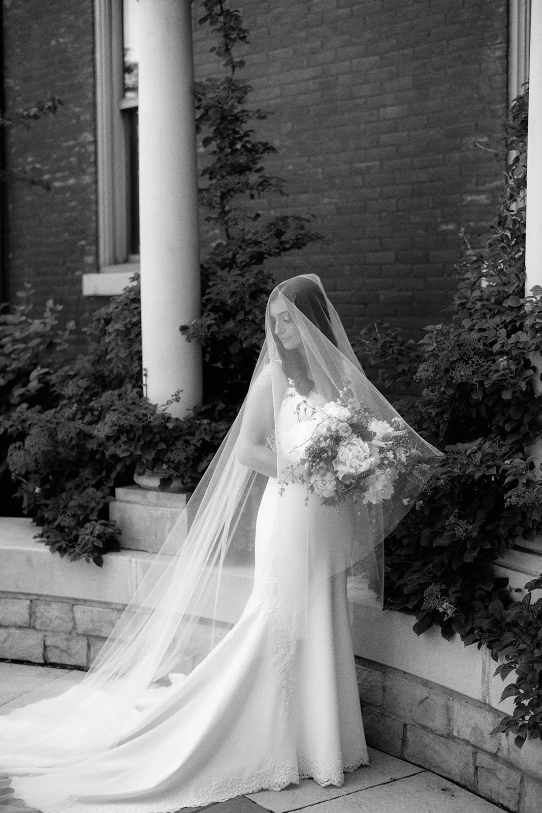 A bride in a wedding dress and veil holding a bouquet, standing outdoors near a brick building with potted plants.