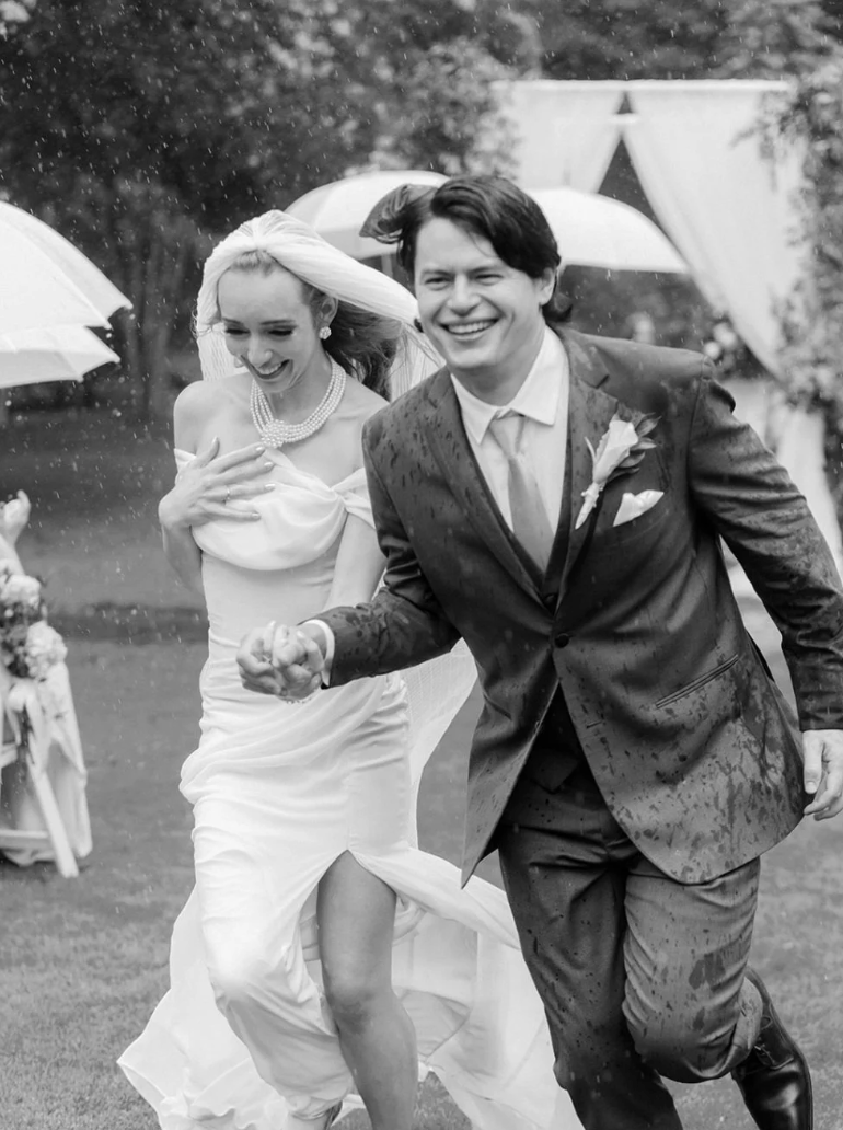 A black-and-white photo of a bride and groom running outdoors, smiling, during their wedding celebration. The bride is wearing a strapless wedding dress and pearl jewelry, while the groom is in a suit with a boutonniere. They are holding hands, and there are umbrellas and decorations in the background.