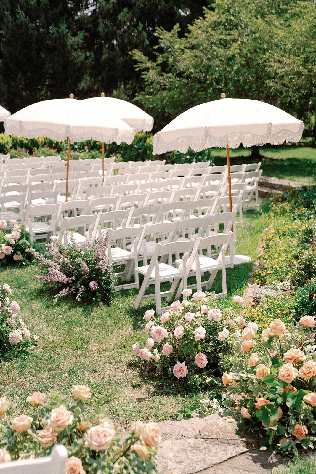 Luxury Outdoor Garden wedding setup in French Park Cincinnati with white chairs arranged in rows, decorated with pink and peach flowers, under large white umbrellas, surrounded by blooming roses and greenery.