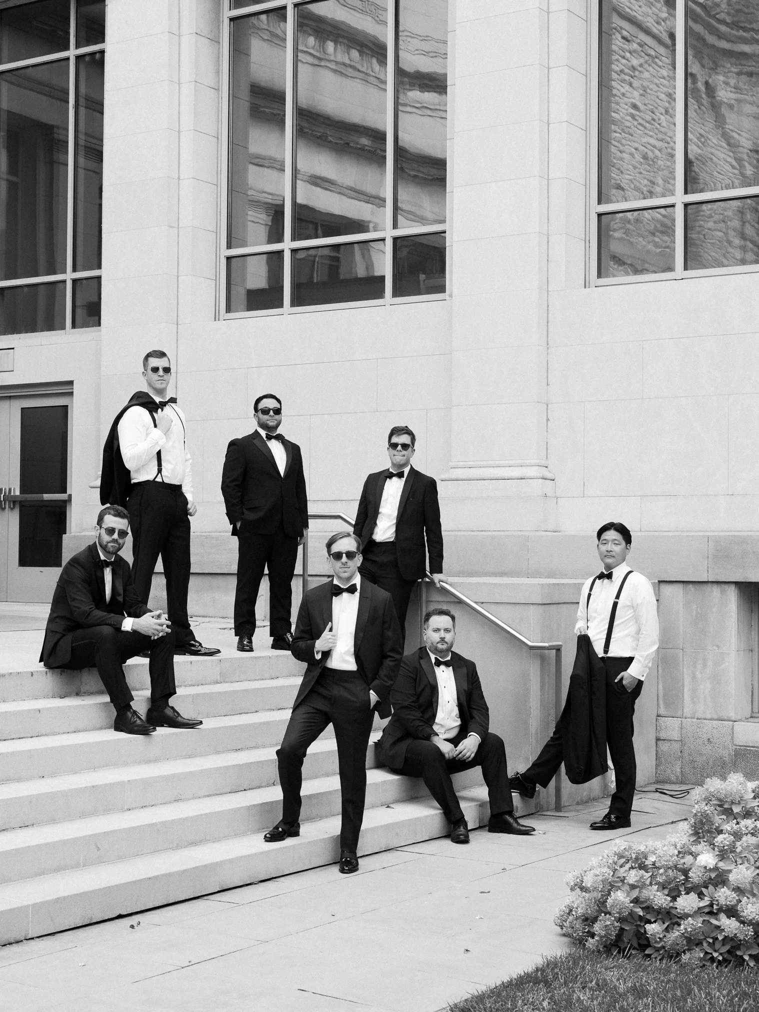 Seven men in tuxedos and sunglasses posing on steps outside a modern building