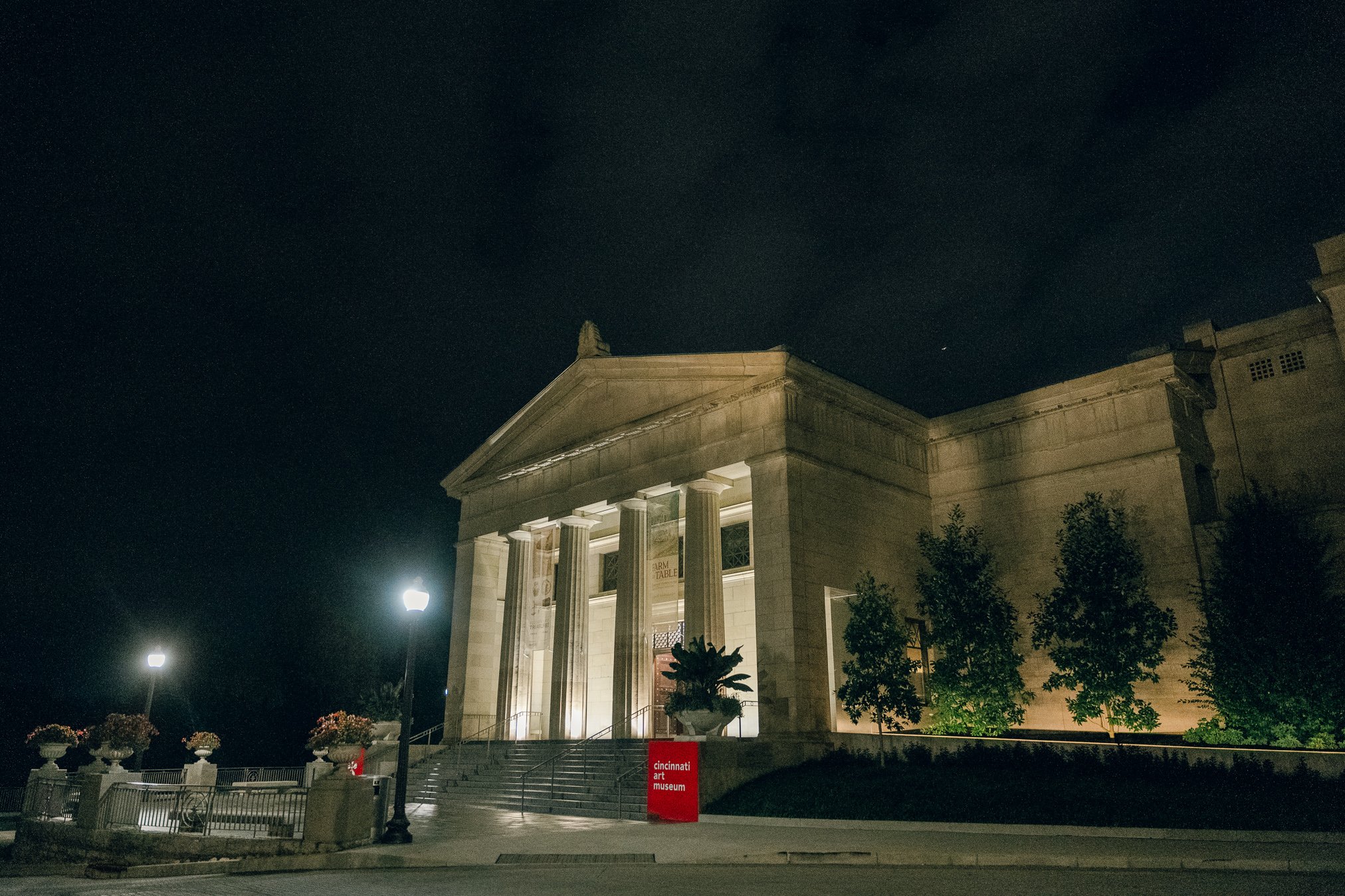 Night view of the Cincinnati Art Museum, a classical-style building with tall columns, steps leading up to the entrance, and trees illuminated by lights.