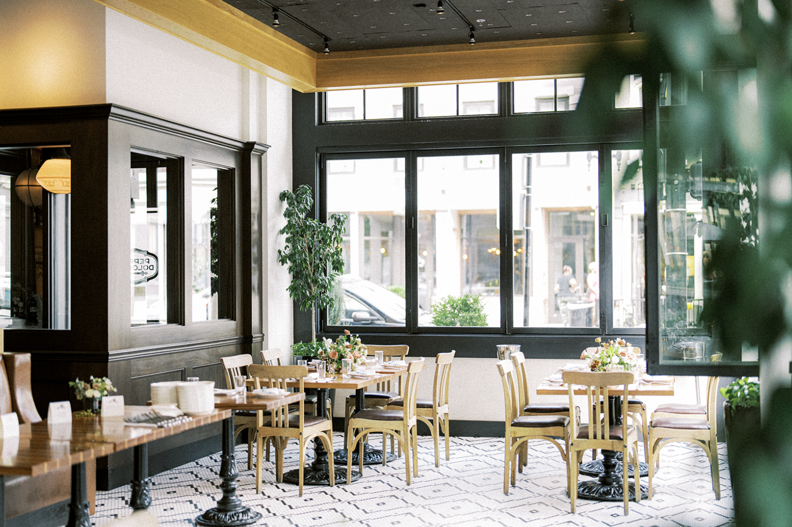 Interior of an elegant restaurant with wooden tables and chairs, large windows, and decorative plants.