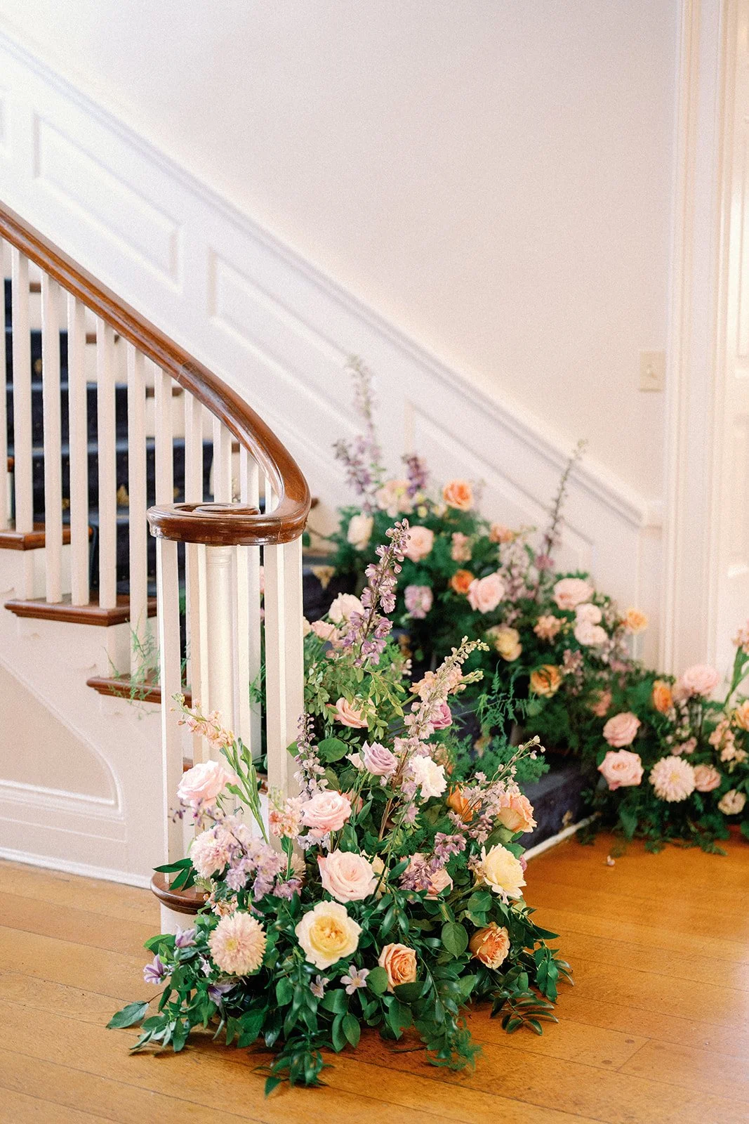 Colorful flower arrangement with pink and peach roses, purple and white blossoms, green foliage, placed along a wooden staircase with white and brown railings, on a hardwood floor inside the French House Cincinnati