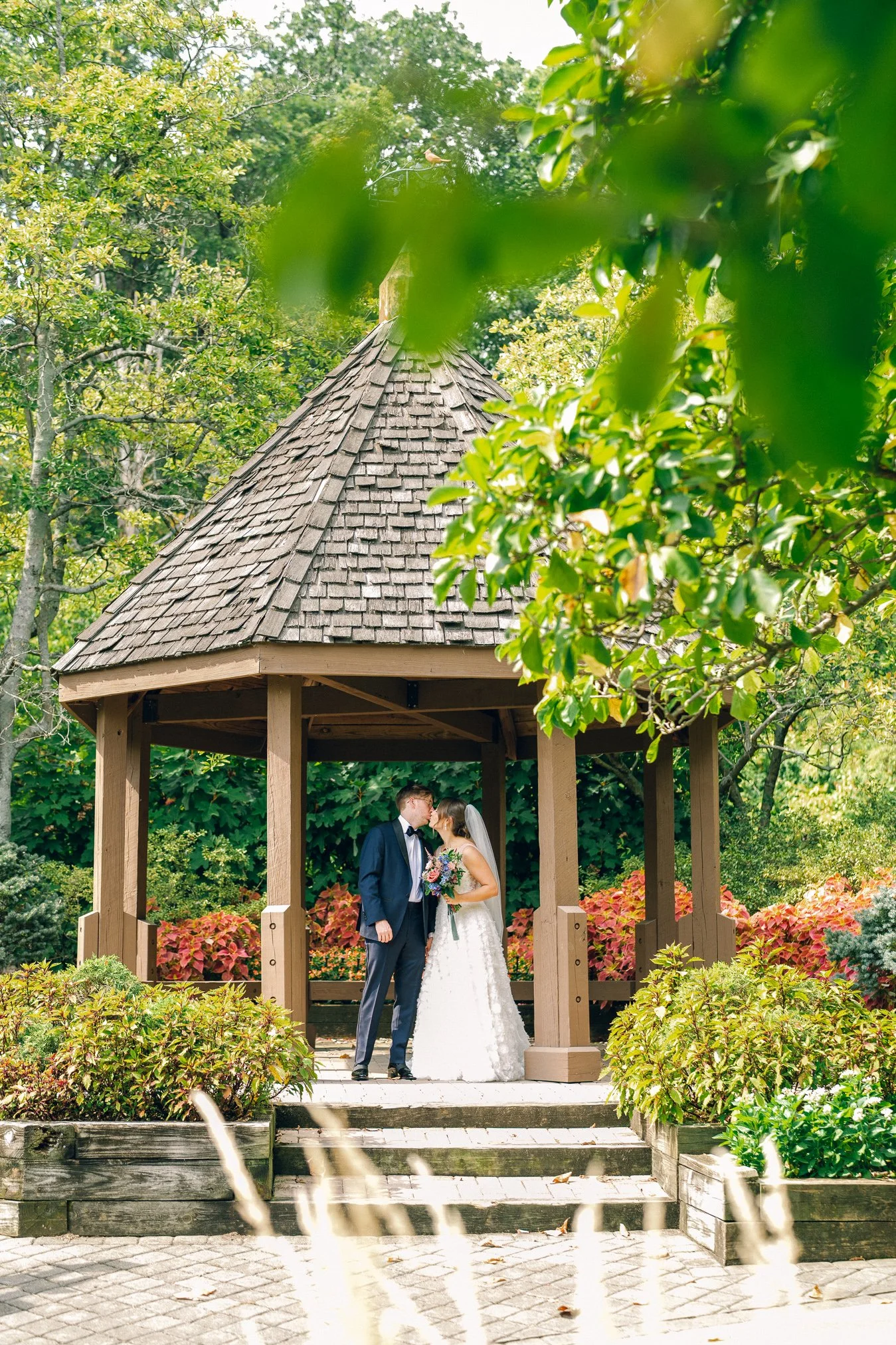 A newlywed couple sharing a kiss under a wooden gazebo in a lush garden.