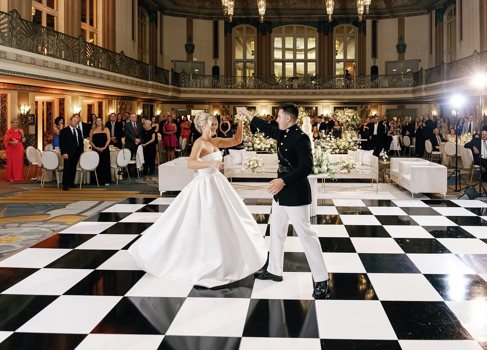 A bride and groom dancing at their wedding reception on a black-and-white checkered dance floor. The bride is in a strapless white wedding gown, and the groom is in a formal military uniform. Guests are watching from tables and the balcony in a luxur