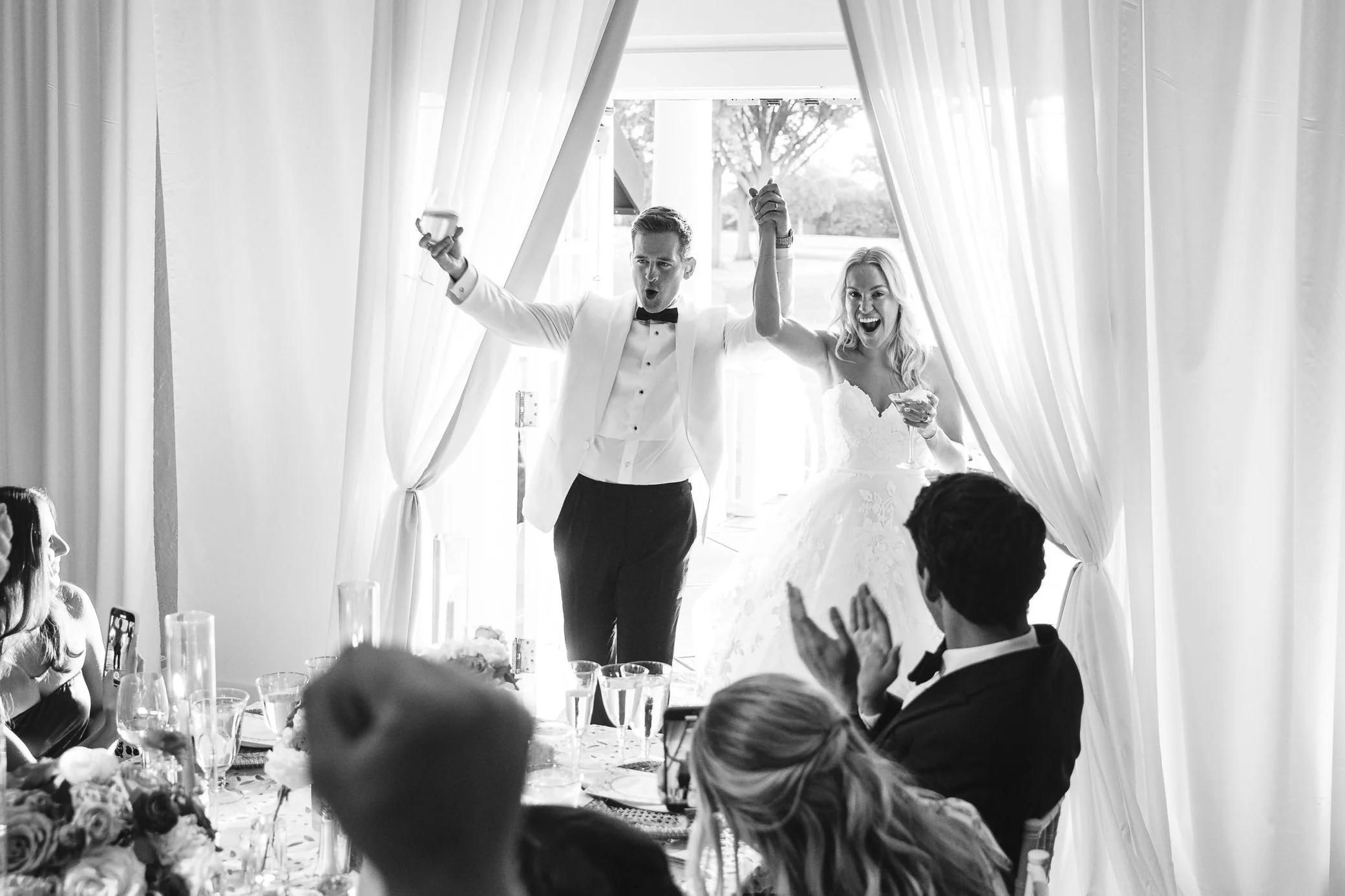 Bride and groom entering a wedding reception, holding hands and celebrating, surrounded by cheering guests.