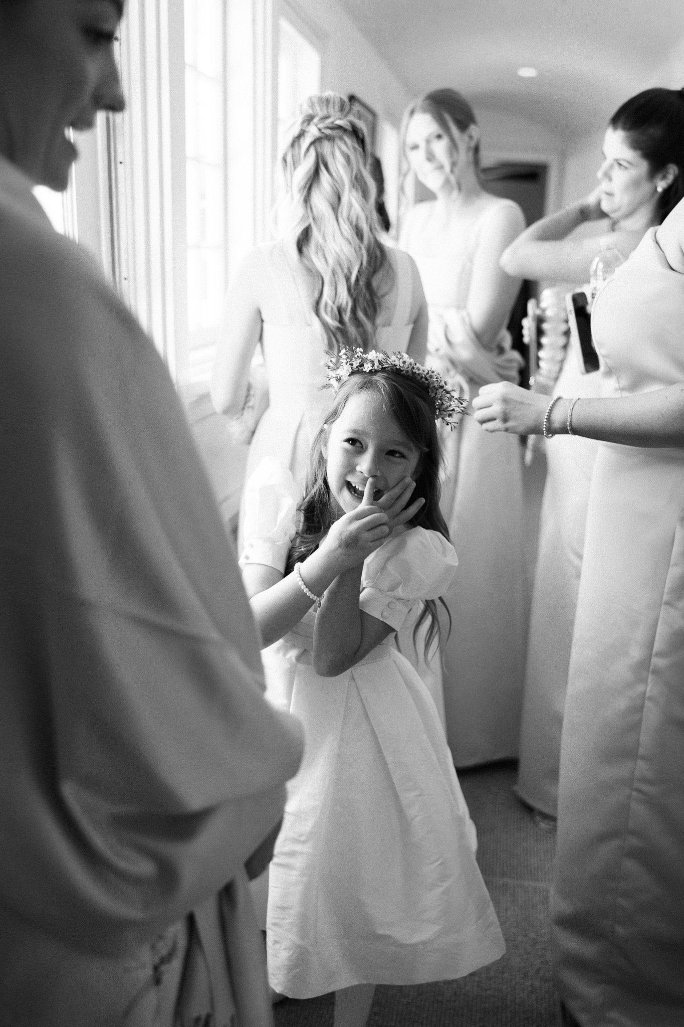 Young girl in a white dress with a flower crown smiling and covering her mouth with her hand, surrounded by women in a bright room.
