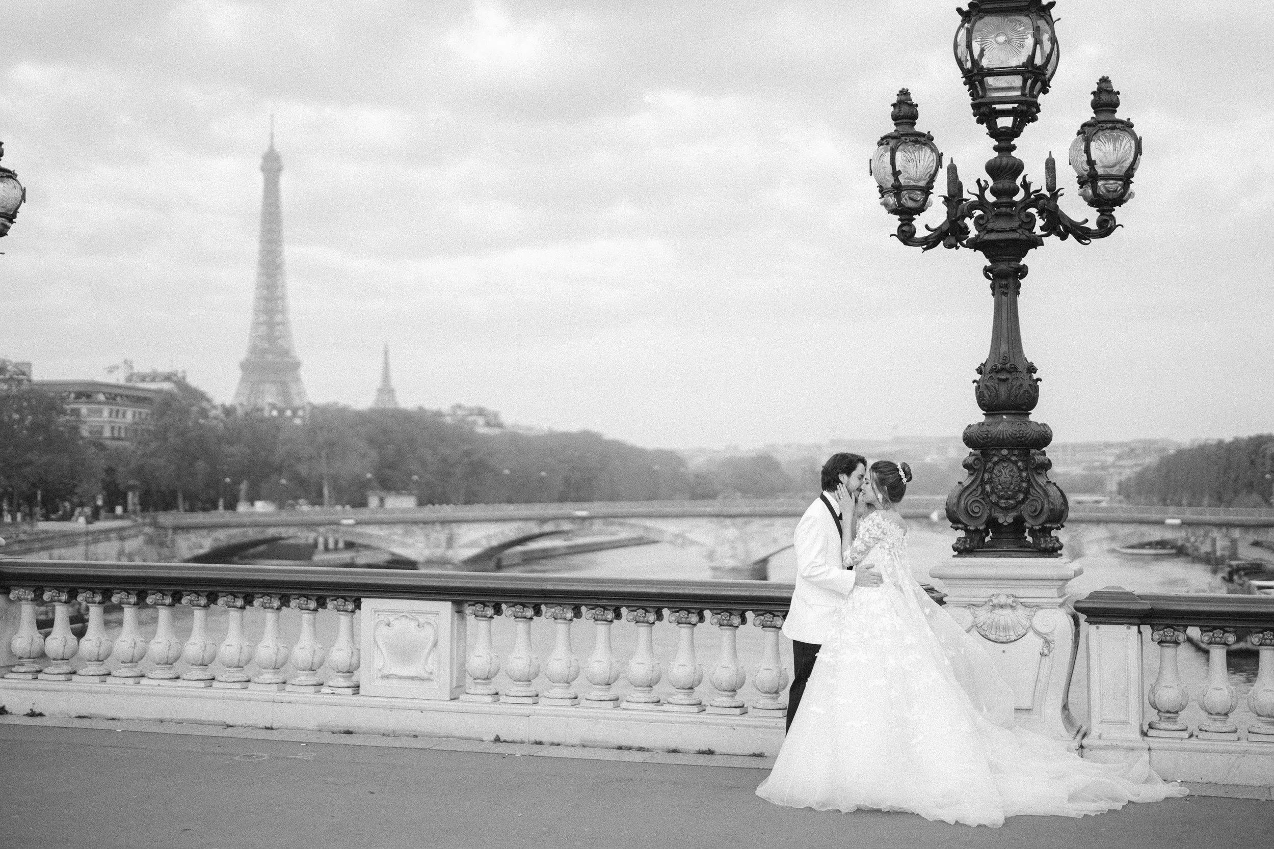 A black and white photo of a newlywed couple kissing on a bridge in Paris with the Eiffel Tower in the background.
