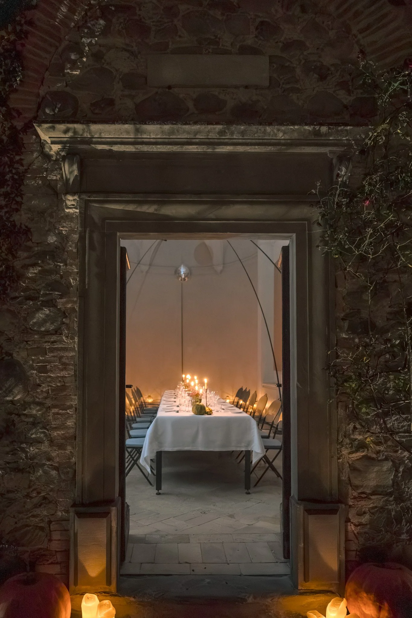 A candlelit dinner table inside a rustic stone building viewed through an open doorway, and candles on the ground outside.
