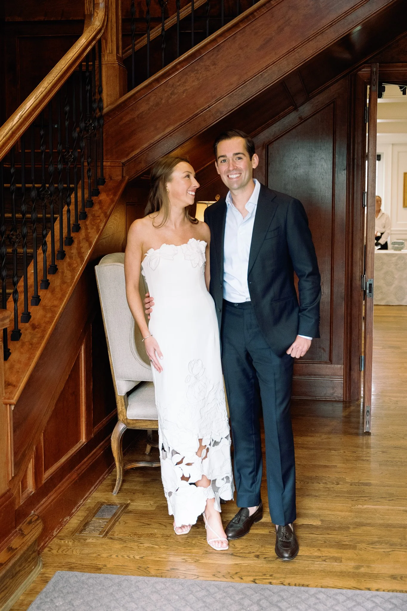 A smiling woman in a white dress and a man in a dark suit pose together indoors under a staircase, with a wooden wall and a doorway behind them.
