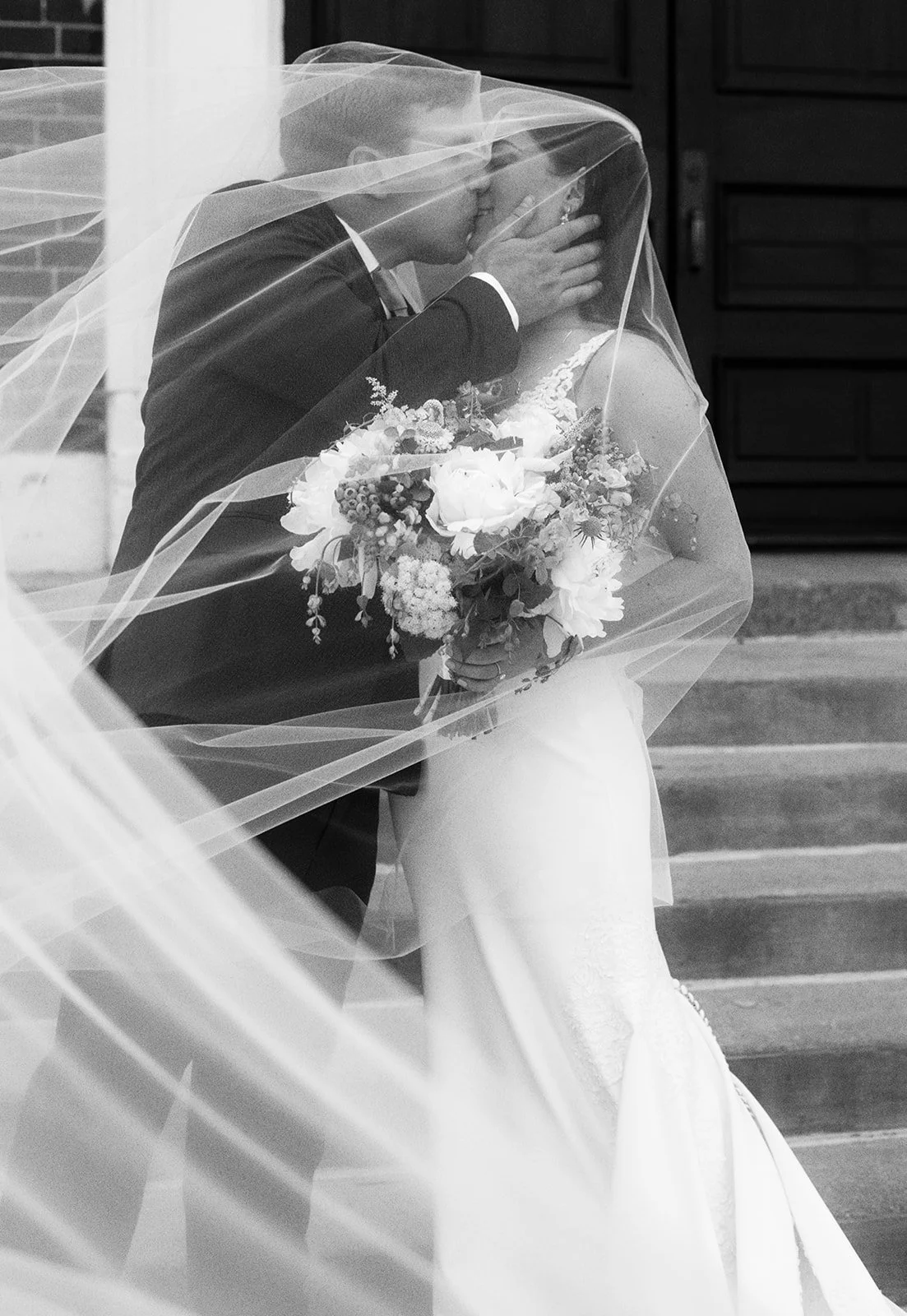 A bride and groom share a kiss under a wedding veil, with the bride holding a bouquet of flowers, on outdoor steps in black and white.
