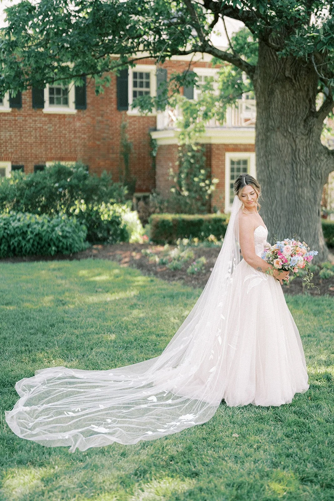 A bride in a white wedding gown with a long veil stands on grass under a large tree, holding a colorful bouquet, in front of the French House at French Park in Amberly Village Cincinnati
