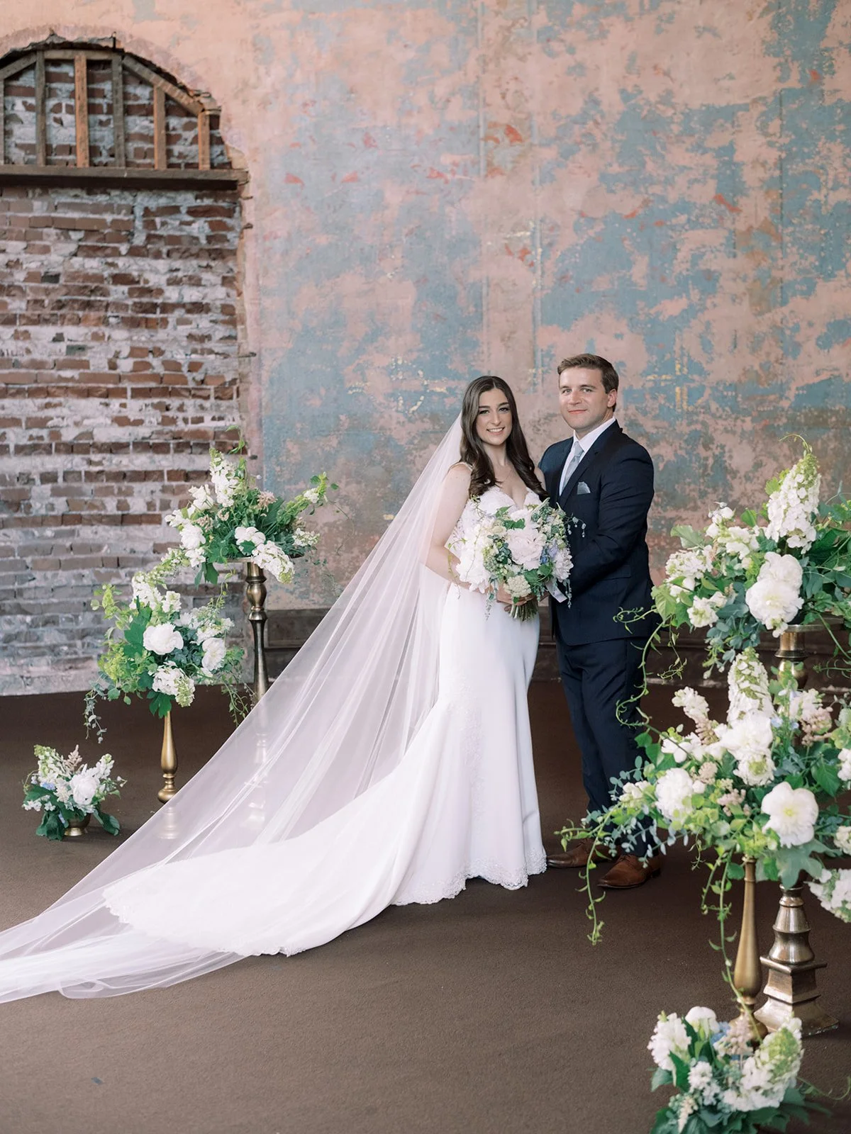 A bride and groom standing together inside a decorated venue, with floral arrangements around them. The bride is wearing a white wedding gown and veil, holding a bouquet, and the groom is dressed in a dark suit. They are smiling and posing for a wedd