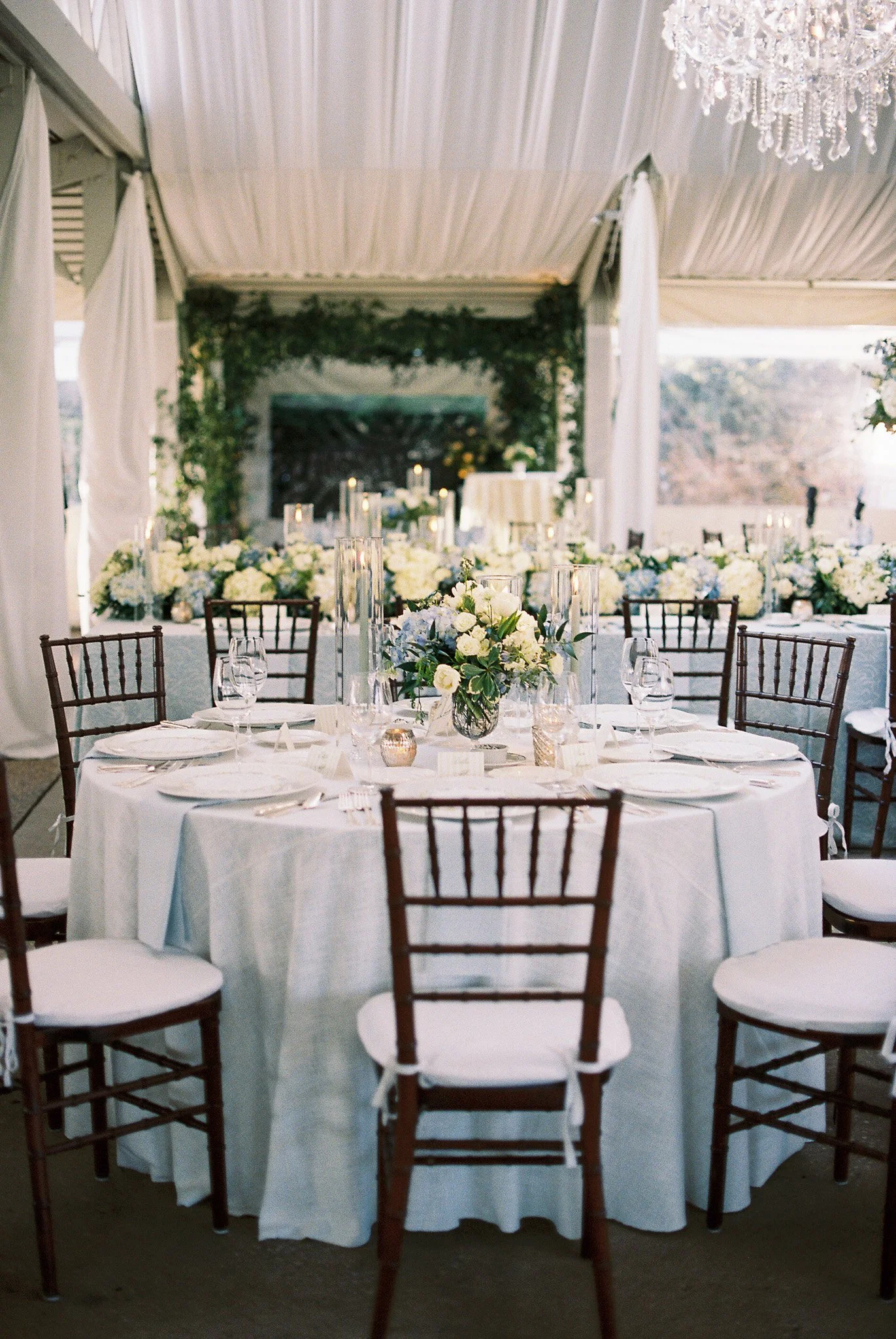 Elegant wedding reception table decorated with white floral arrangements, tall candle holders, and glassware inside a decorated tent with draped fabric and a chandelier.