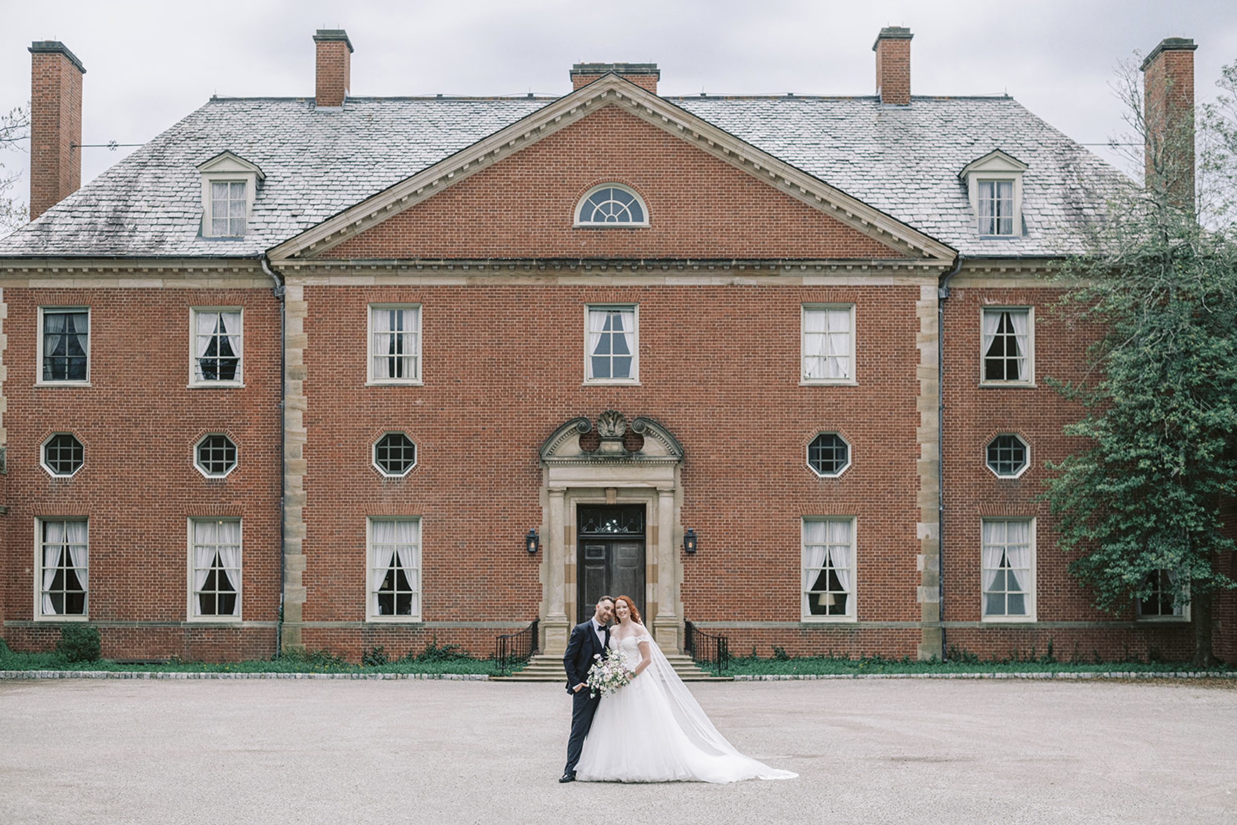 Historic brick building with large windows, decorative stone carvings, and a flowering shrub in the foreground.