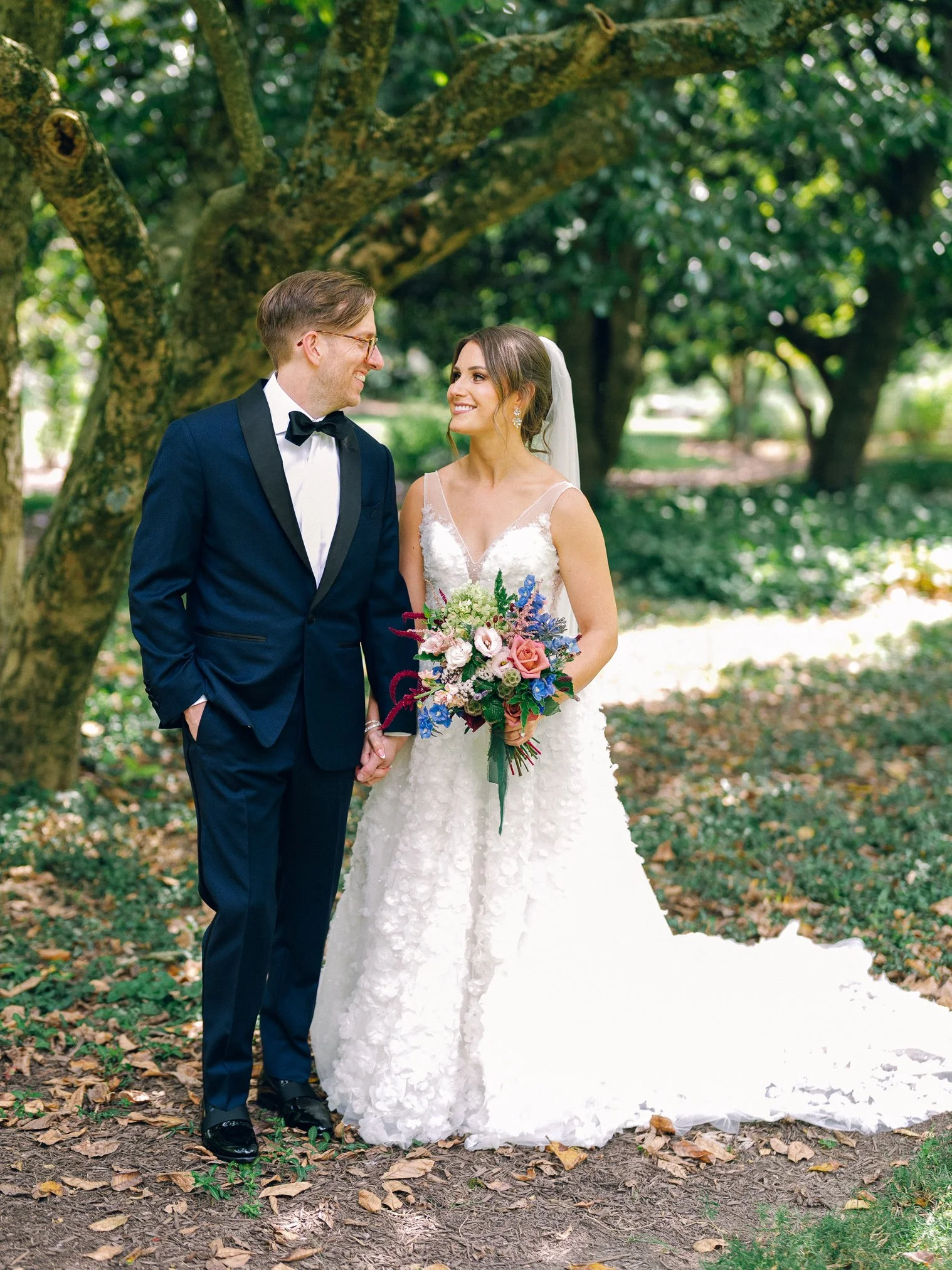 A bride and groom holding hands and smiling at each other outdoors near a large tree with green foliage, with the bride holding a colorful bouquet of flowers, both dressed in wedding attire.