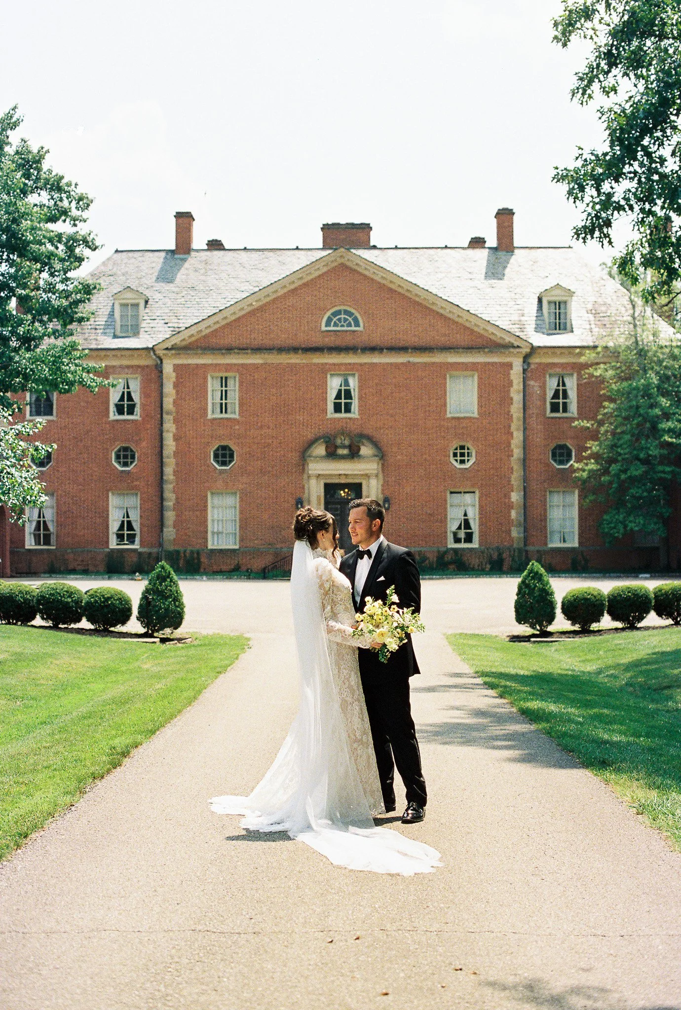 A bride and groom in wedding attire standing on a pathway in front of a large brick mansion, holding hands and facing each other.