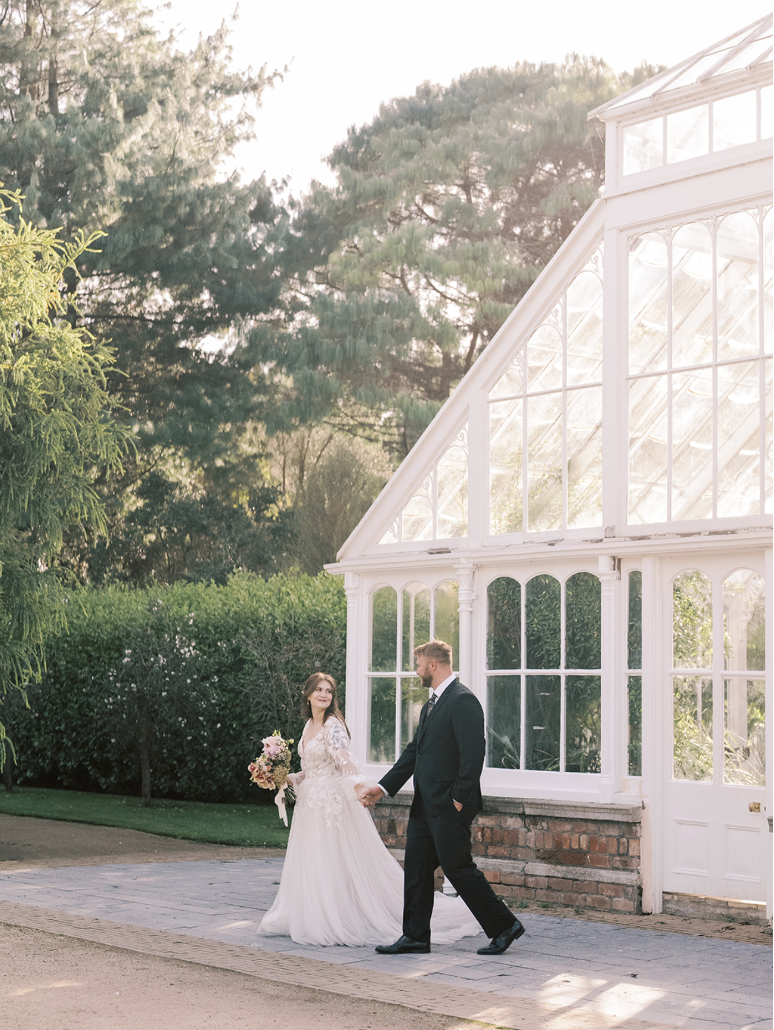 Bride and groom walking hand in hand outside a greenhouse, with trees and bushes in the background, during daytime.