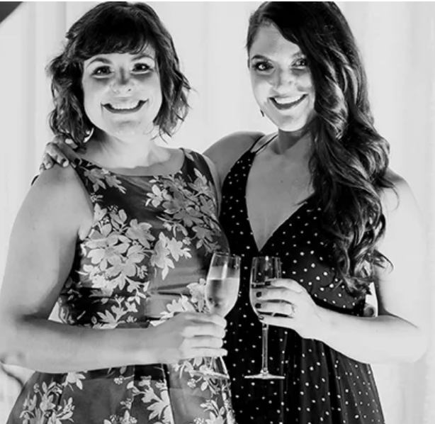 Two women in elegant dresses smiling and holding champagne glasses at a formal event.