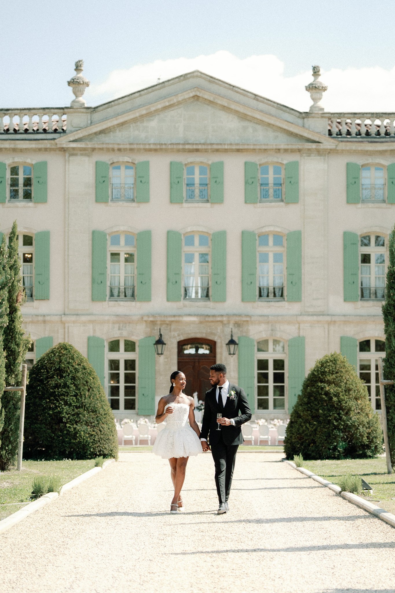 A newlywed couple walking hand in hand on a gravel path outside a grand, historic building with green shutters, clear sky and manicured bushes.