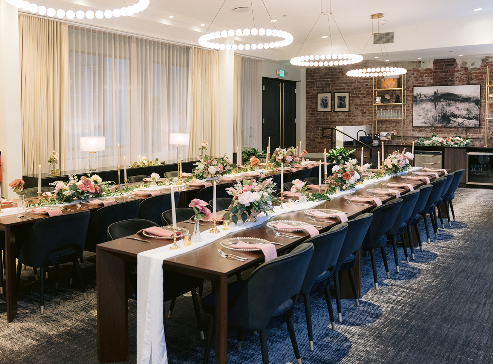 Elegantly decorated dining table set for a formal event, featuring floral centerpieces, pink napkins, gold candle holders, and modern chandeliers in an upscale venue with exposed brick walls and large windows.