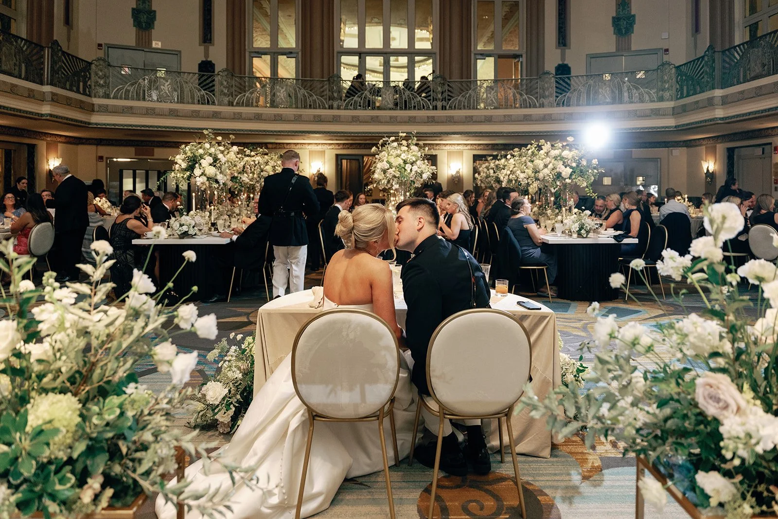 A wedding reception in a grand hall with a bride and groom seated at a table, surrounded by floral arrangements and guests dining at round tables, with a balcony and large windows in the background.