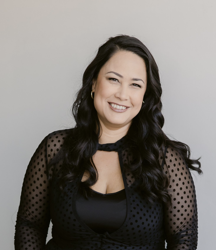 Smiling woman with long dark hair wearing a black polka dot blouse against a neutral background.