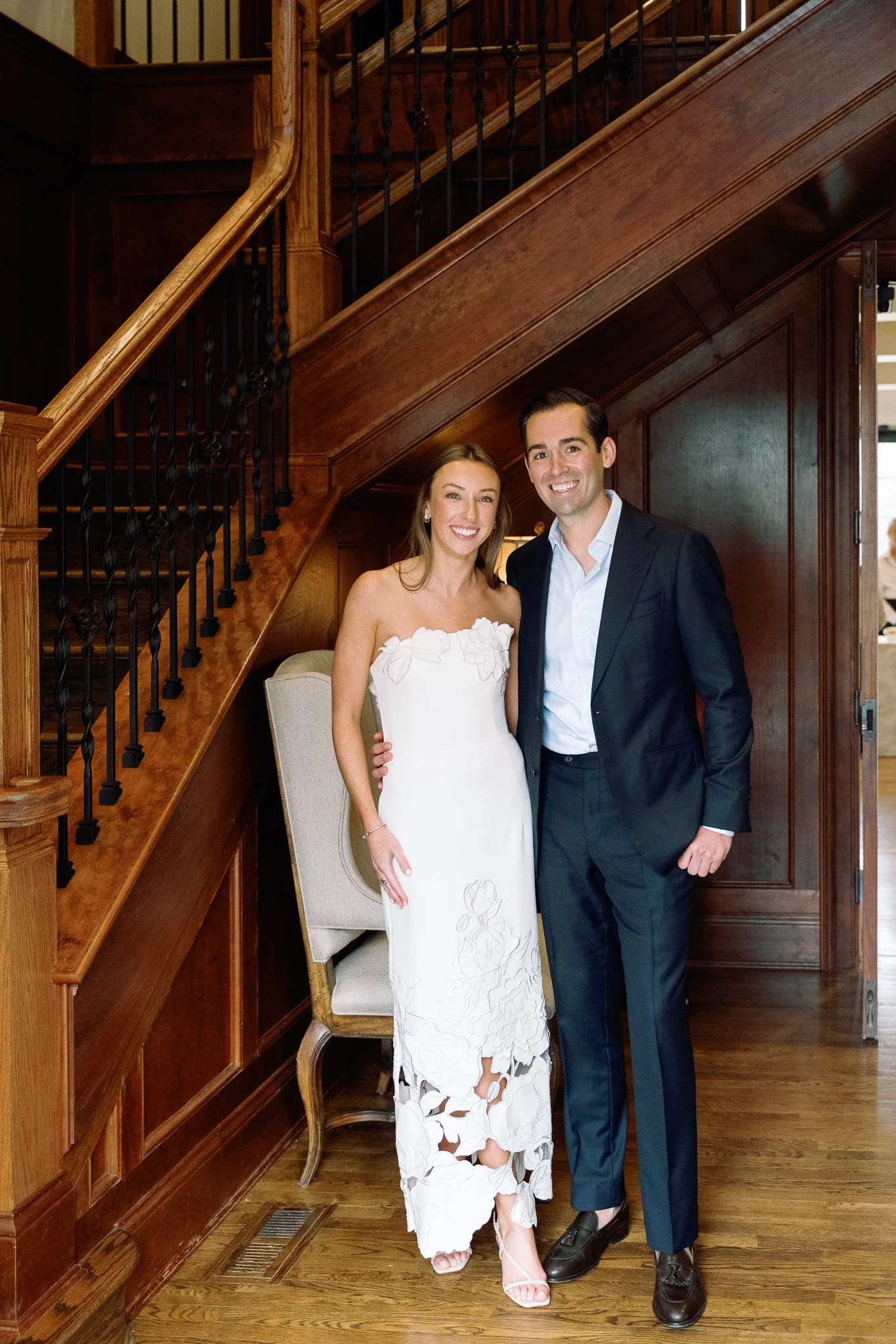 A couple dressed in formal attire, standing indoors under a wooden staircase, smiling at the camera.