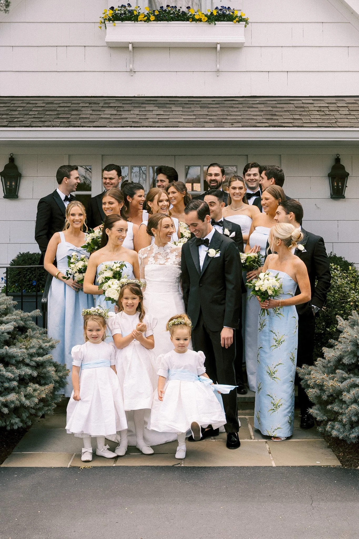 A group of people, including a bride and groom, posing for a wedding photo in front of a white house with shrubs and flowers. The bride wears a lace wedding dress, and the groom is in a black tuxedo. Several women in light blue dresses hold bouquets,