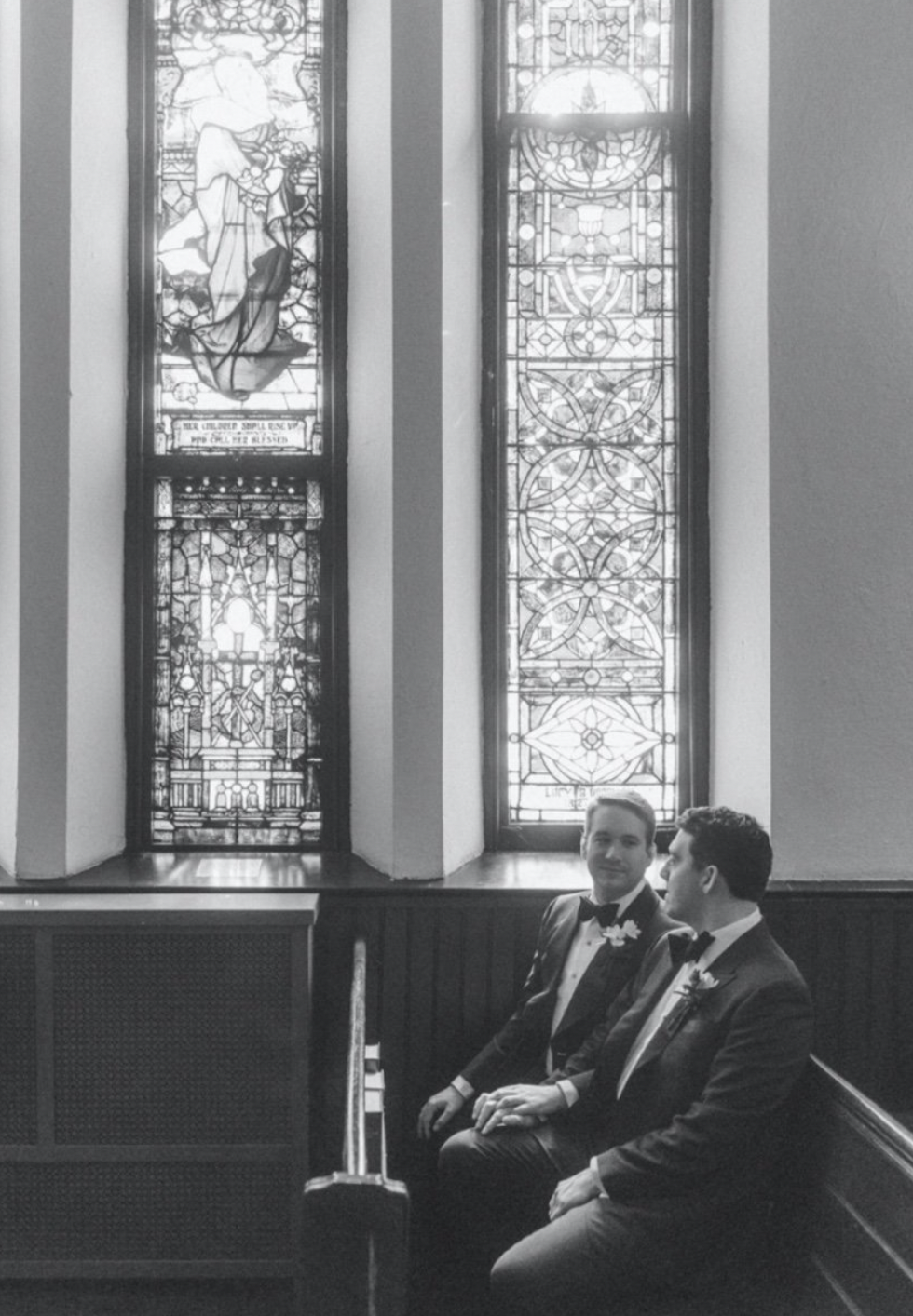 Two men in tuxedos sitting in pews inside a church, with stained glass windows behind them.