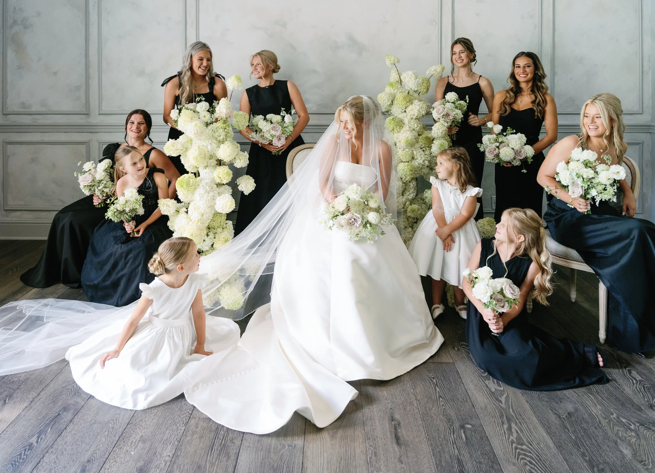 Bride with bridesmaids and flower girls in formal attire, holding bouquets, in an elegant setting with floral decorations.
