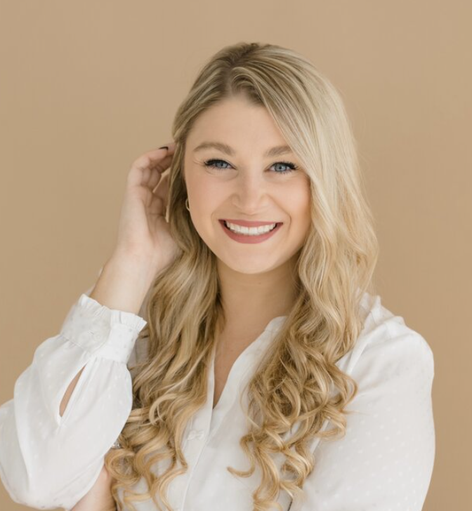 Woman with long blonde hair smiling, wearing a white blouse, against a beige background.