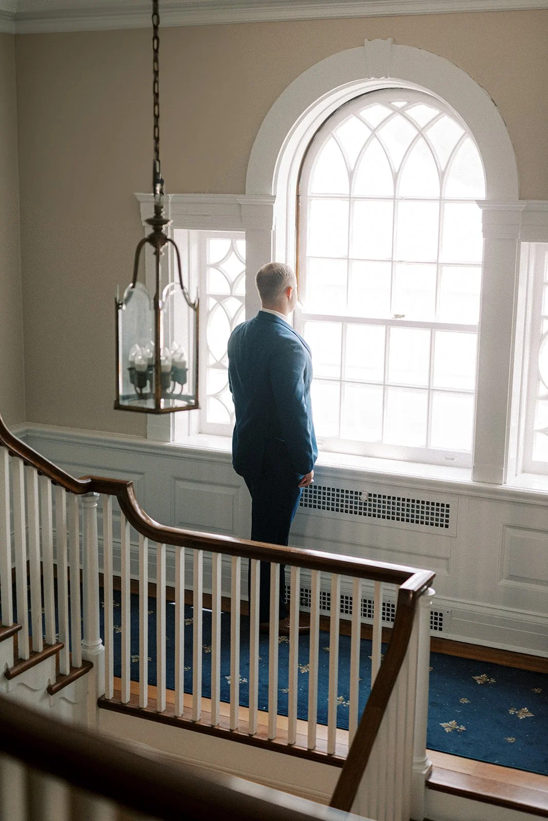 A man in a blue suit standing by a large window in a well-lit, elegant interior with white walls, wooden staircase railing, and a blue carpet inside the French House Cincinnati