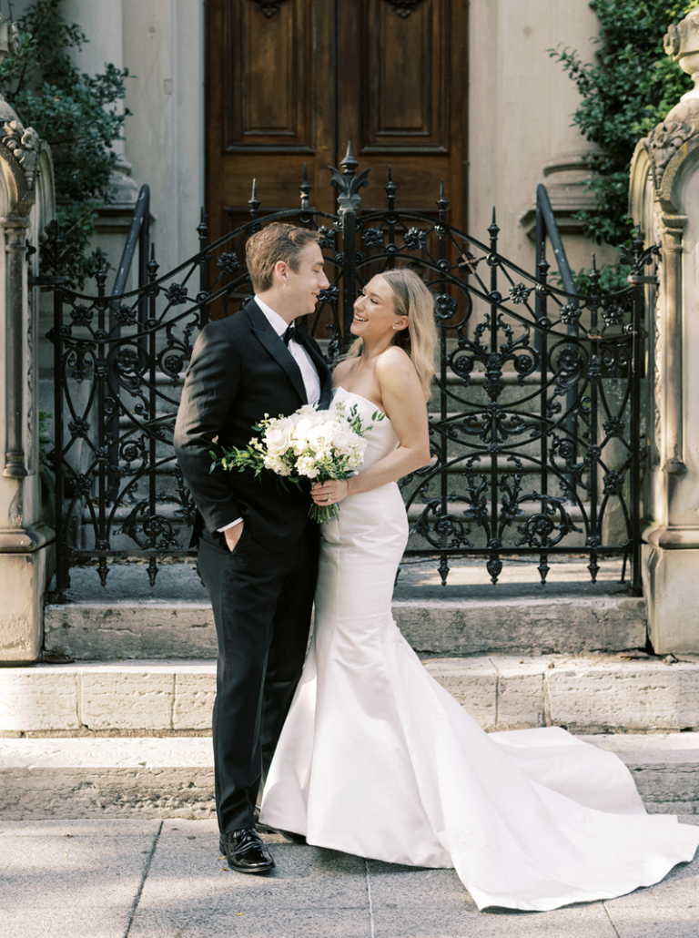 A newlywed couple stands on the steps in front of a wrought-iron gate and stone columns, smiling and looking at each other. The bride wears a strapless white wedding gown and holds a bouquet of white flowers. The groom wears a black tuxedo with a white shirt and bow tie.