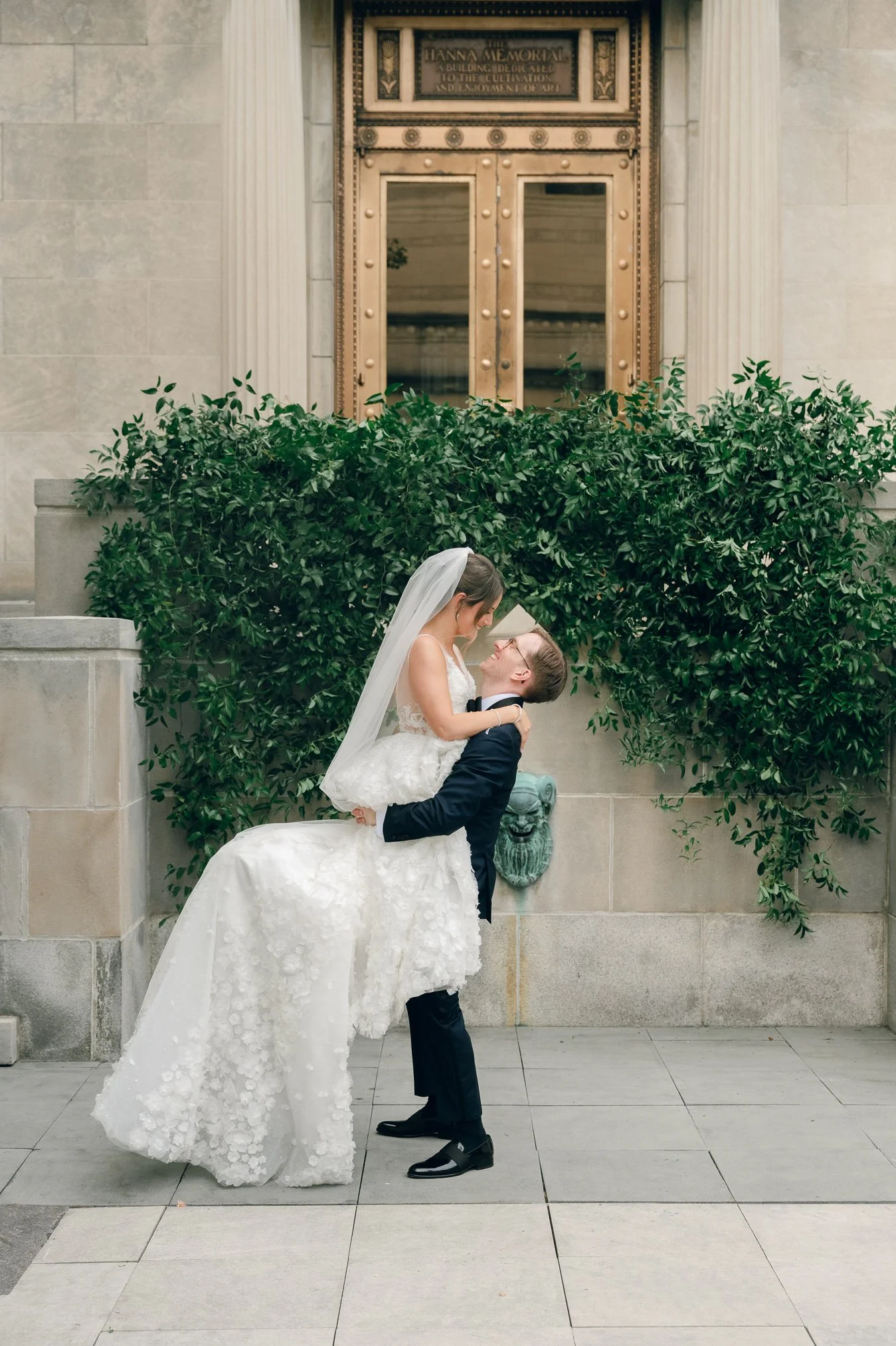 A bride and groom on their wedding day, with the groom lifting the bride as they gaze at each other, standing in front of a large green bush and ornate building entrance.