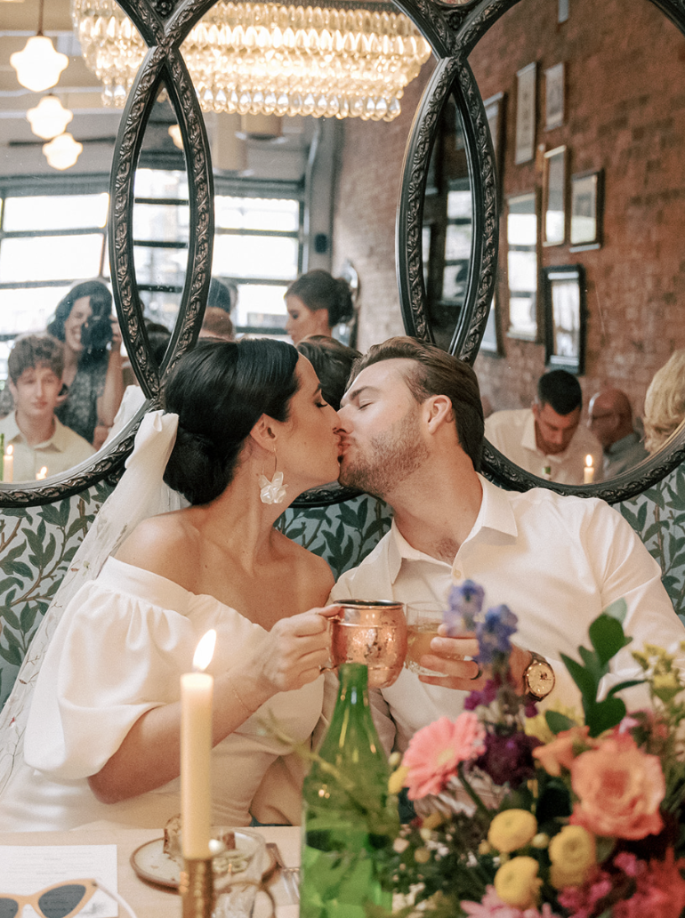 A couple sharing a kiss at a wedding reception, visible through a decorative mirror. The bride wears a white dress and a veil, while the groom is in a white shirt. They sit at a table with colorful flowers, candles, and drinks, in a decorated indoor setting.