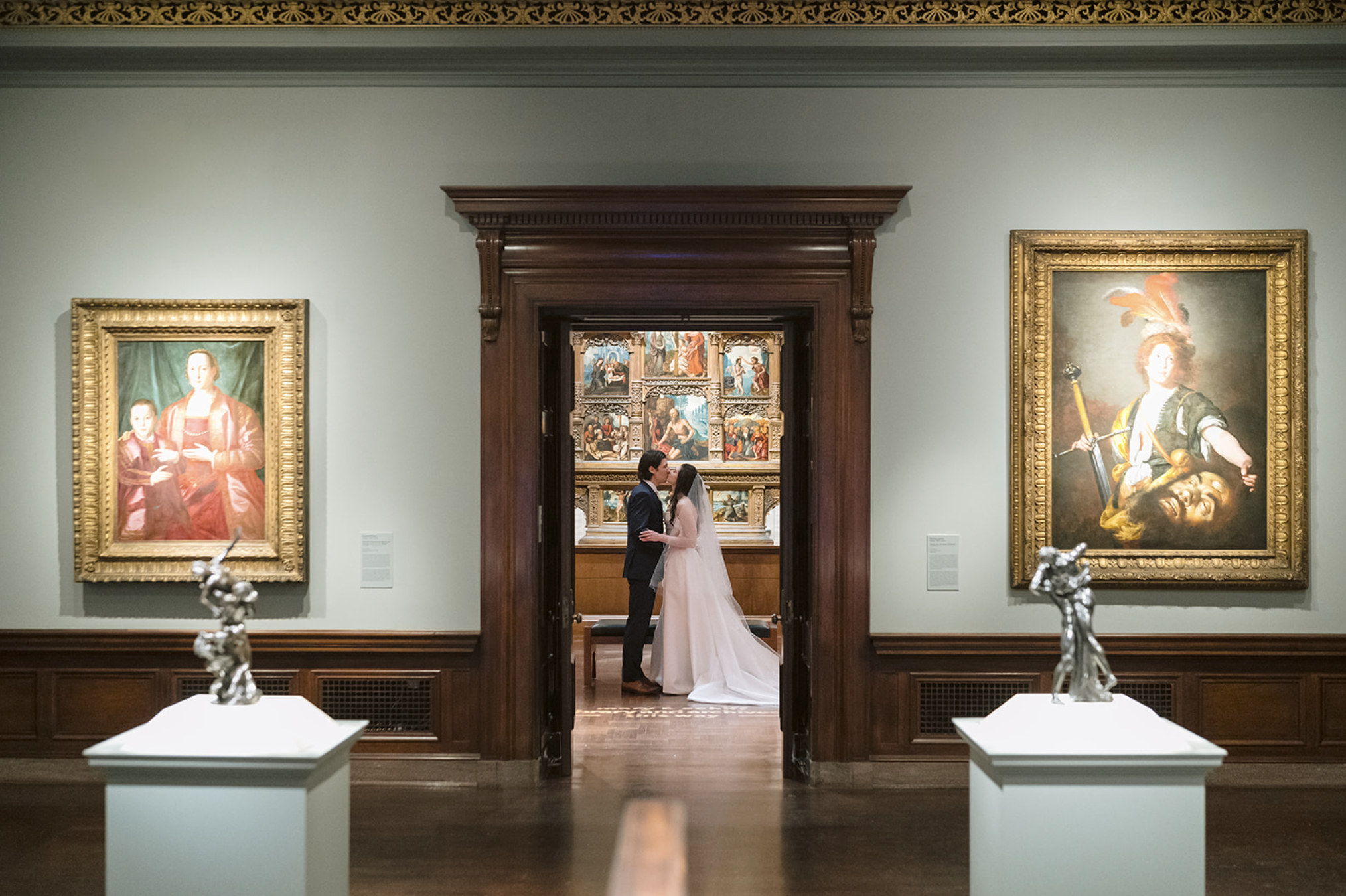 A bride and groom stand kissing in a hallway of an art gallery, surrounded by framed paintings and sculptures.