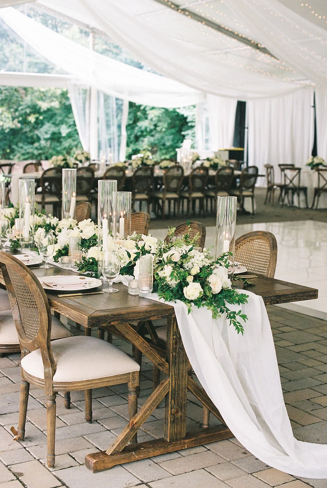 Tented Wedding reception at Peterloon Estate in Cincinnati. Table decorated with white flowers, candles in tall glass holders, and a white table runner, set inside a tent with white drapes and string lights.