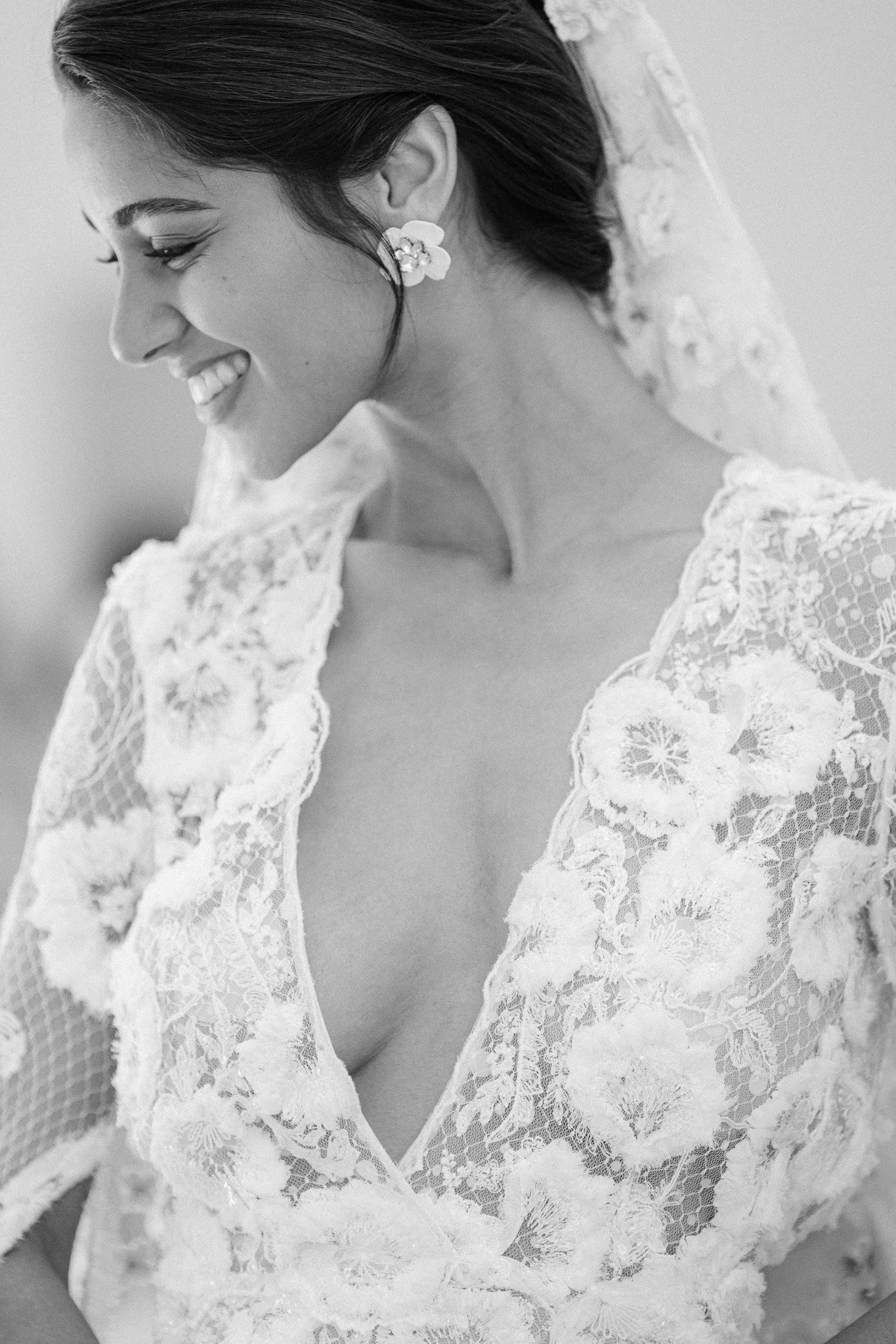 A smiling woman in a wedding dress with lace floral details and a deep V neckline, her hair styled back, wearing floral earrings.