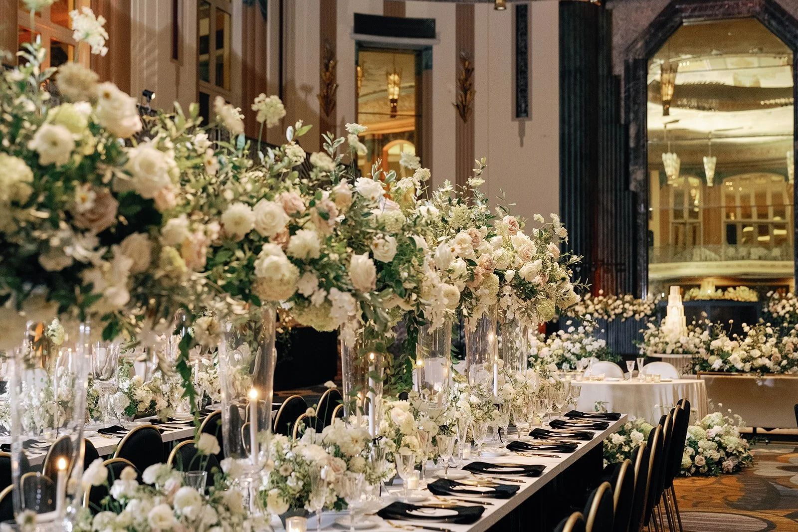 A wedding reception at the Hall of Mirrors decorated with tall floral centerpieces featuring white and blush roses, carnations, and greenery on a long table with black napkins and glassware, in an elegant ballroom with ornate walls and large windows.