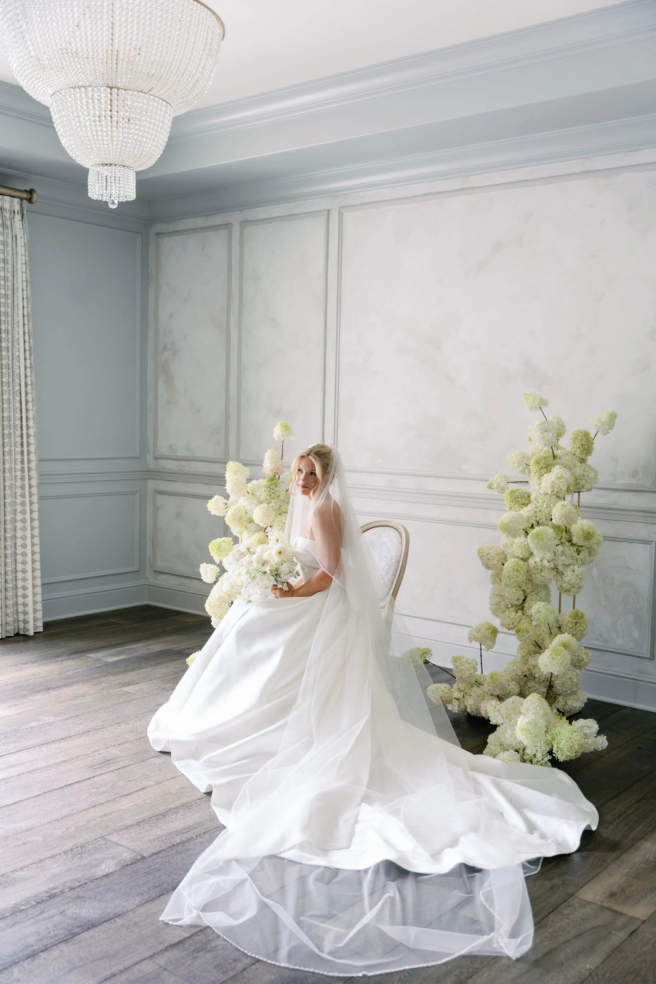 Bride in a white wedding gown and veil sitting on a chair in a decorated room with large white floral arrangements and a chandelier.