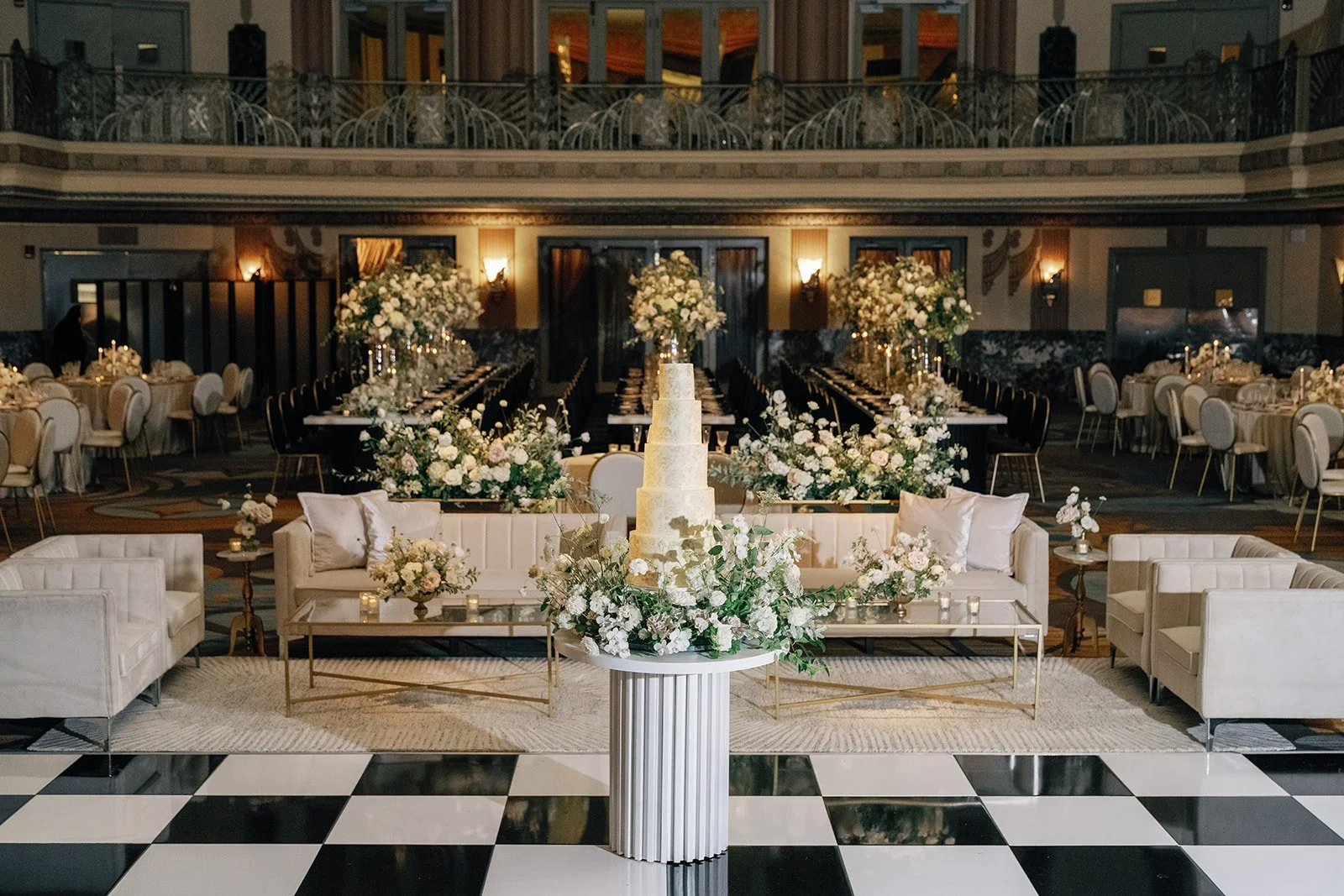 Elegant wedding reception hall with a tiered white wedding cake on a white pedestal surrounded by white floral arrangements, ornate tables with chairs, and a mezzanine balcony in the background.