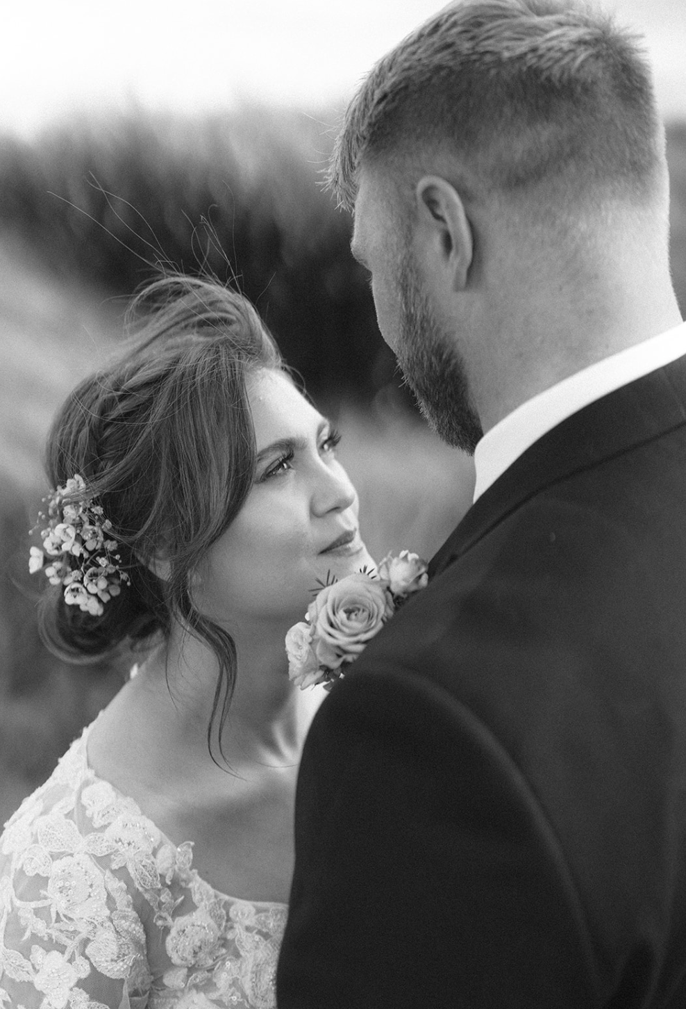 Black and white photo of a bride and groom facing each other, with the groom holding a boutonniere near the bride's face. The bride has flowers in her hair and is wearing a lace dress, while the groom is in a suit.