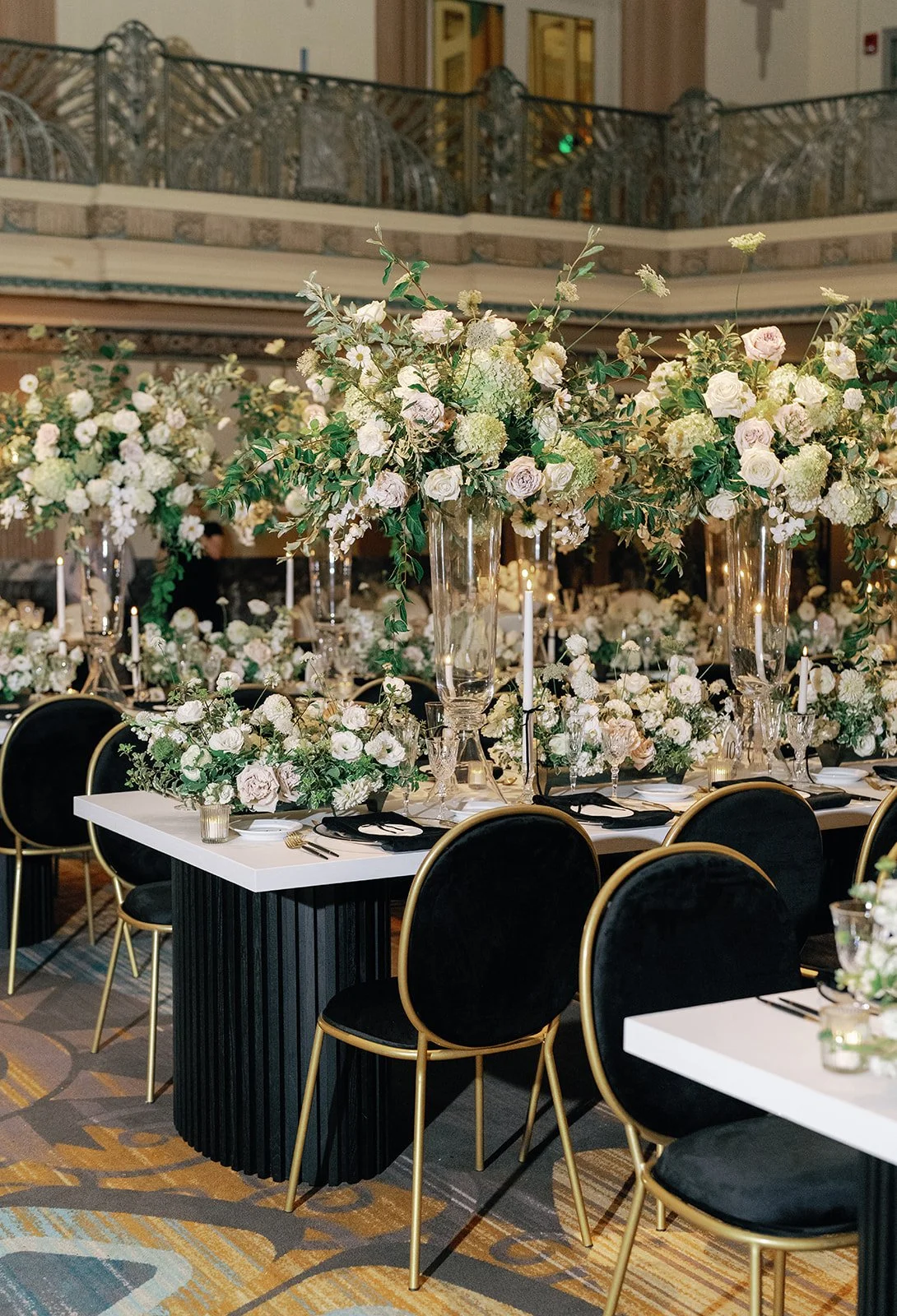 Elegant banquet table decorated with large floral arrangements of white and blush flowers, green foliage, tall glass vases, and black and gold chairs in a grand, ornate hall.