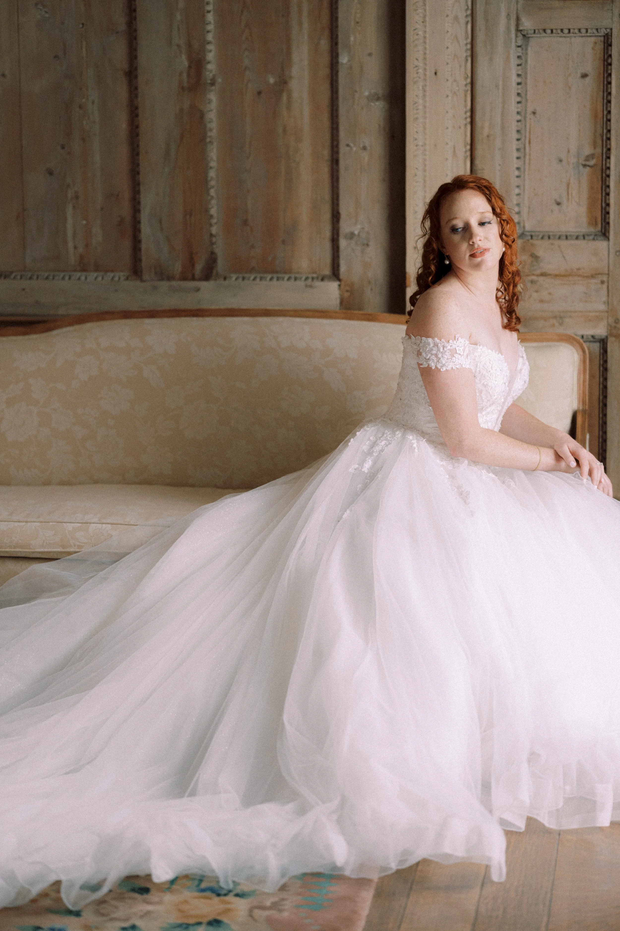 A woman wearing a white wedding gown with off-the-shoulder lace details, sitting on a vintage beige sofa in a rustic wooden room.