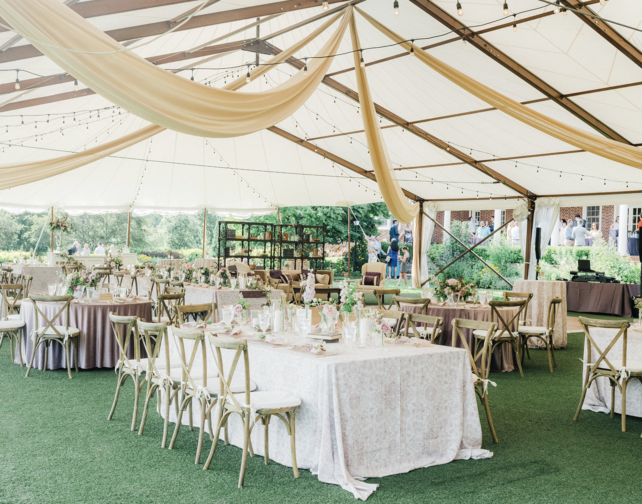 Outdoor wedding reception setup under a tent with elegant lighting, draped fabric, decorated tables and chairs, and floral centerpieces on grass.