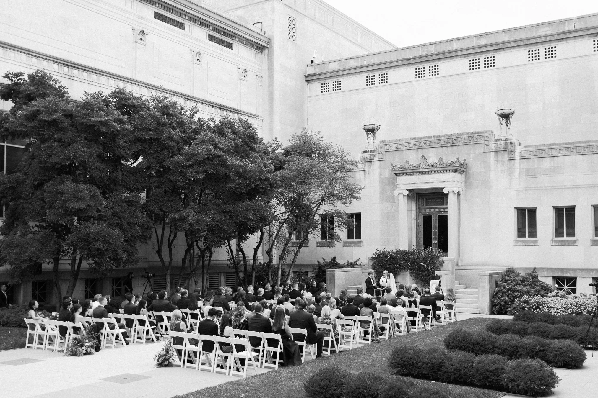A black and white photo of an outdoor wedding ceremony with guests seated in rows of white chairs facing an altar where the couple and officiant stand, surrounded by trees and a large building in the background.