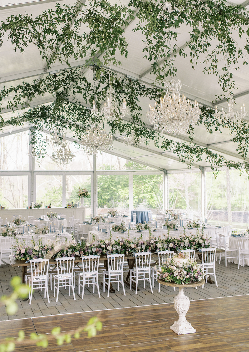 Elegant wedding or event reception inside a bright tent with chandeliers and green hanging foliage, decorated with floral arrangements and tables set for a celebration.